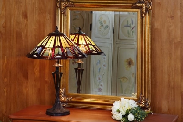 Applique lampe Tiffany et miroir doré au cadre orné sur une commode en bois, bouquet de fleurs blanches posés à côté.