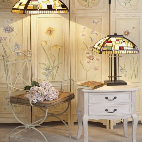 Lampe Tiffany au damier coloré sur une table de chevet blanche près d’un paravent floral et d’un fauteuil en métal peint