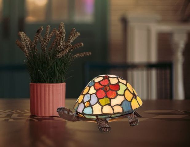 Lampe Tiffany en forme de tortue avec un abat-jour en vitrail coloré posée sur une table, près d’un bouquet de fleurs dans